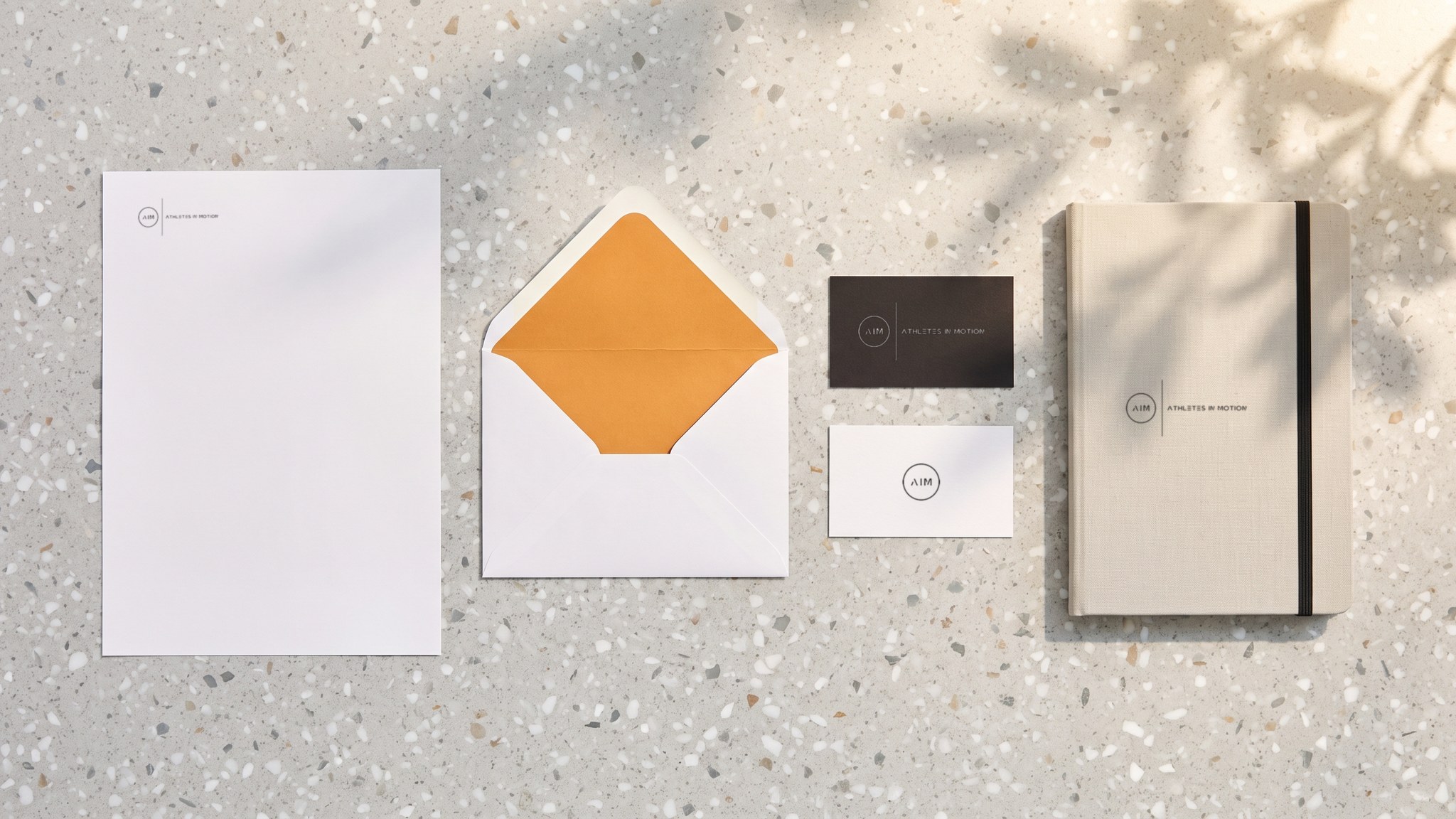 A render seen from above of the AIM letterhead, an envelope with an orange liner, a black card with the white lockup, a white card with the mark and a stone notebook, on terrazzo