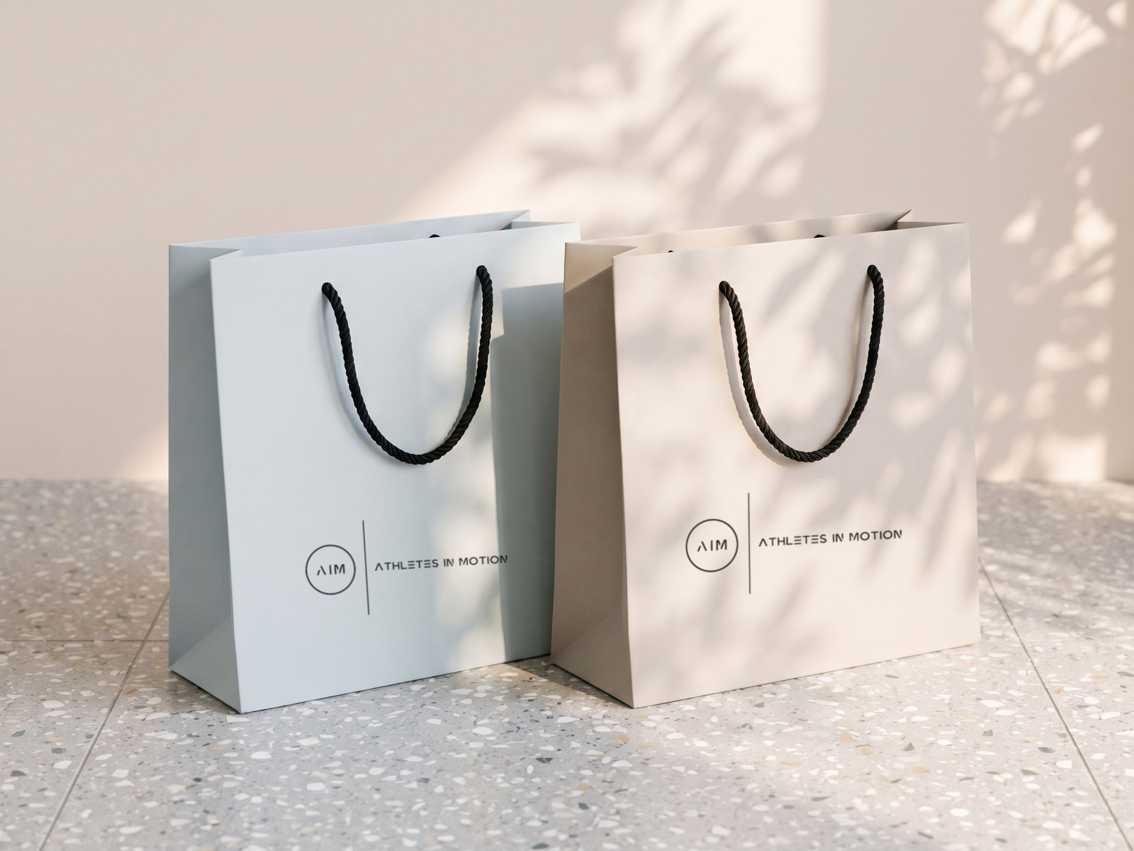A render of two paper bags, pale blue and stone, with black rope handles and the AIM lockup, on terrazzo in leaf shadow