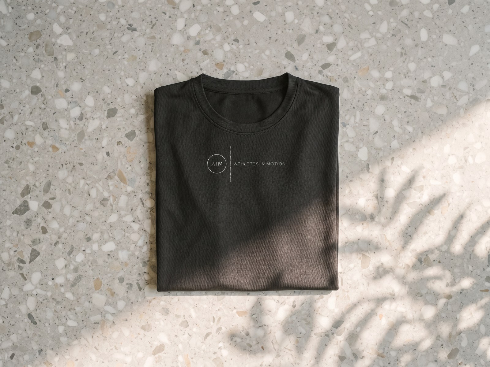 A render of a near-black training t-shirt folded square on terrazzo, ATHLETES IN MOTION and the AIM mark in white across the chest