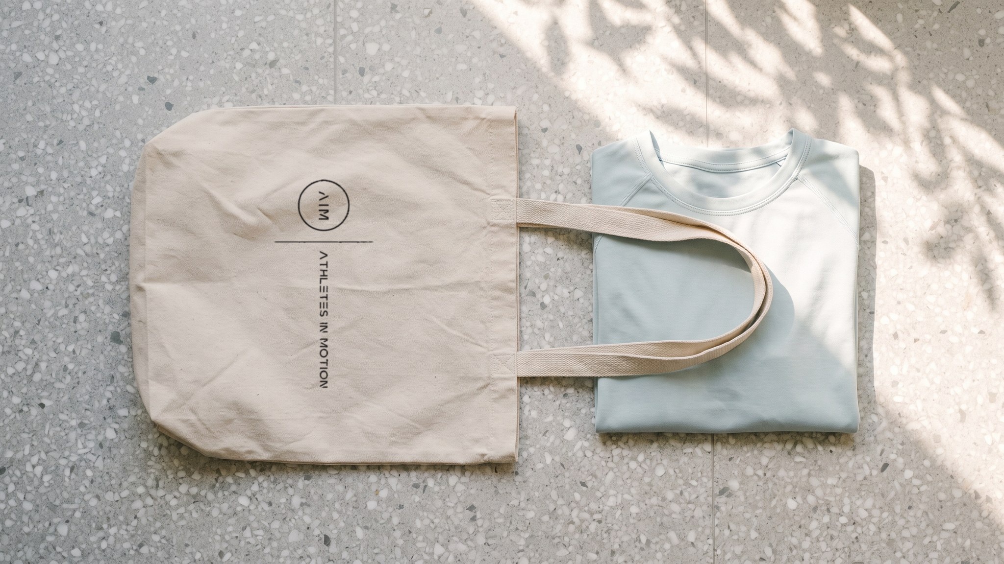 A render seen from above of a canvas tote with the AIM lockup beside a folded pale blue top, on terrazzo