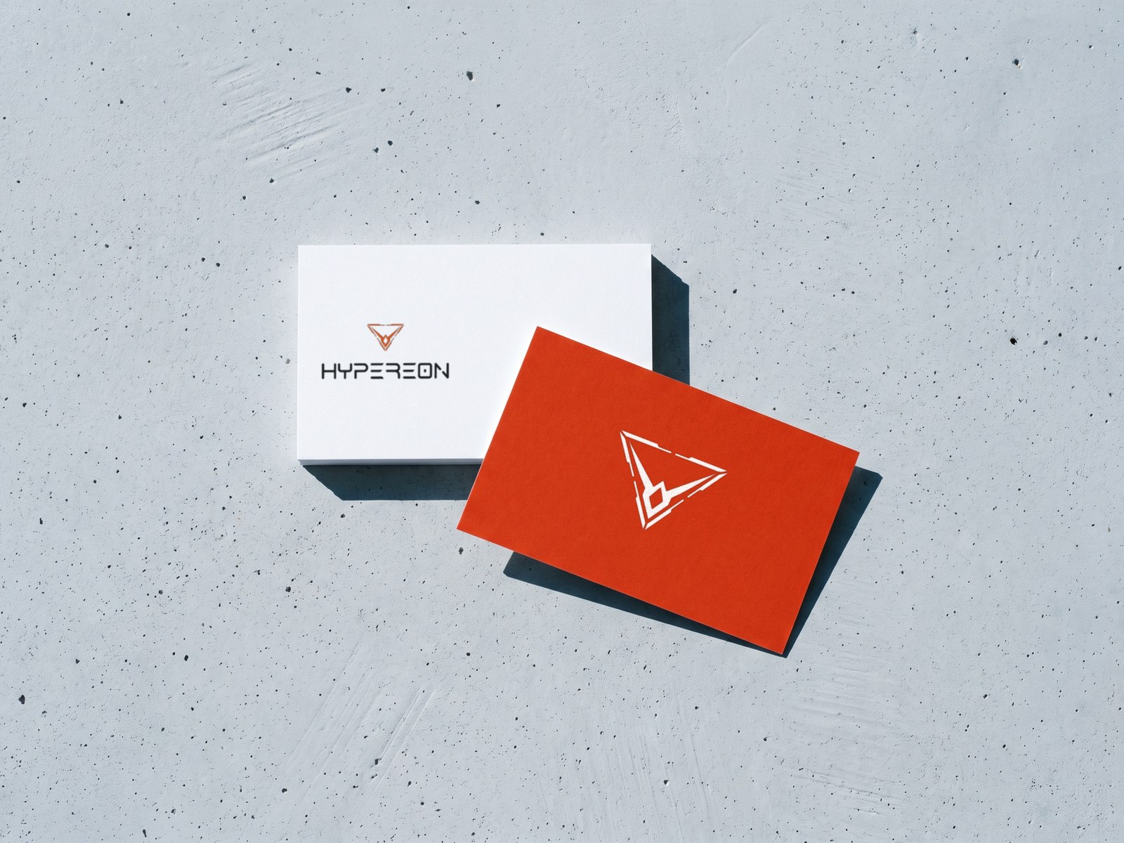 A render of two Hypereon business cards on grey concrete in hard sun: a white card with the orange mark over the dark wordmark, and an orange card with the mark in white
