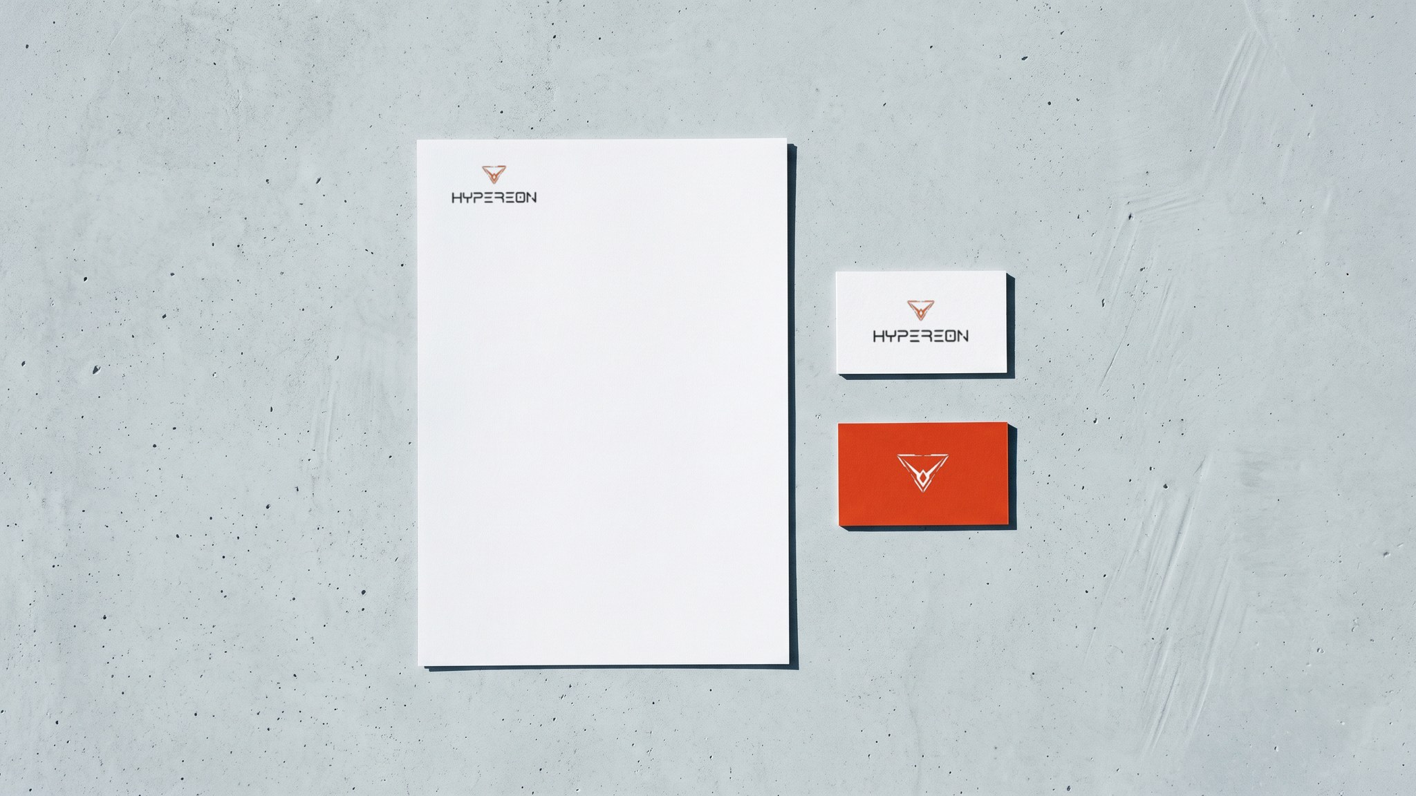 A render seen from above of a white Hypereon letterhead with the logo at its head, a white card with the logo and an orange card with the white mark, on grey concrete