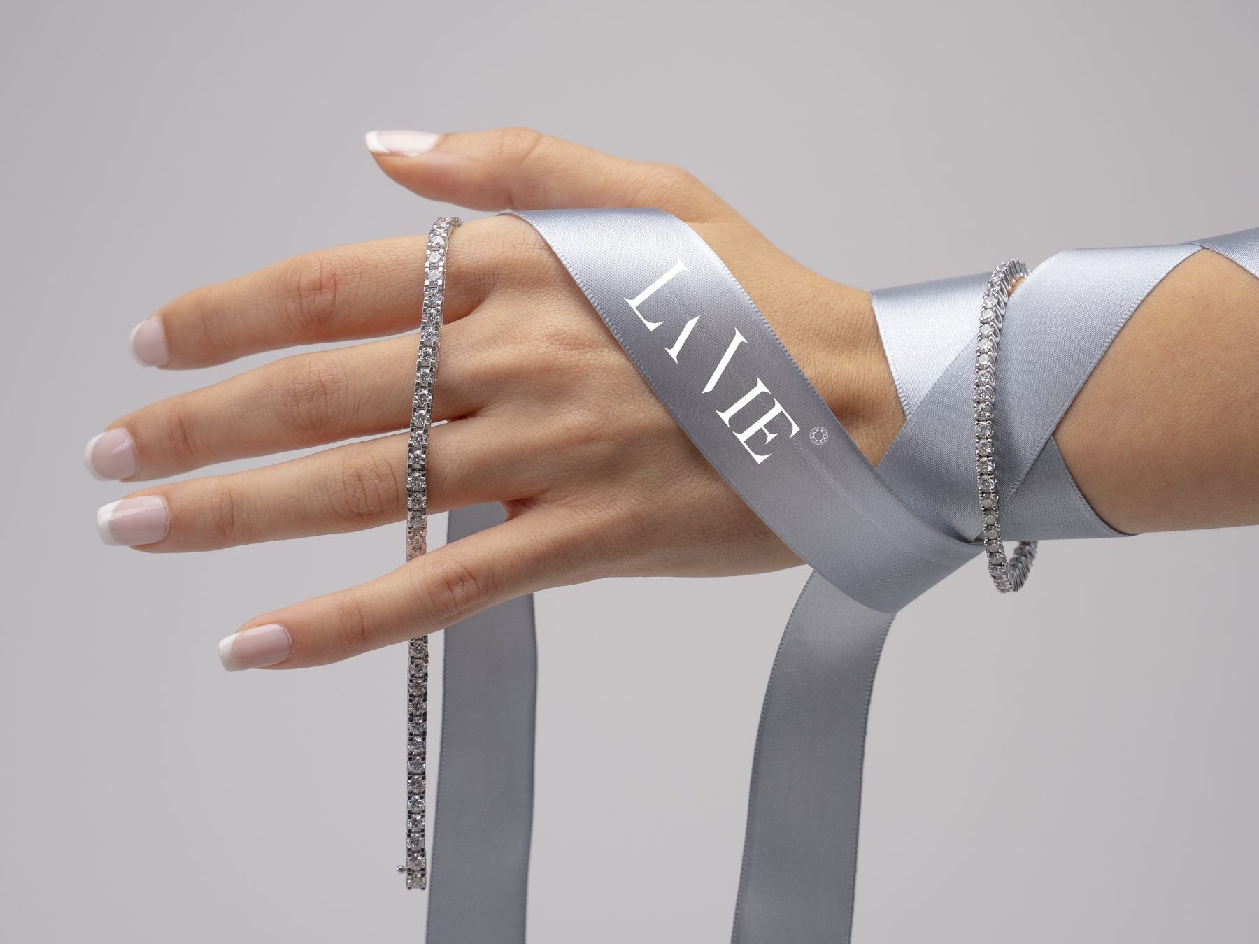 A satin ribbon printed with the La Vie wordmark crossing the back of a hand with two bracelets