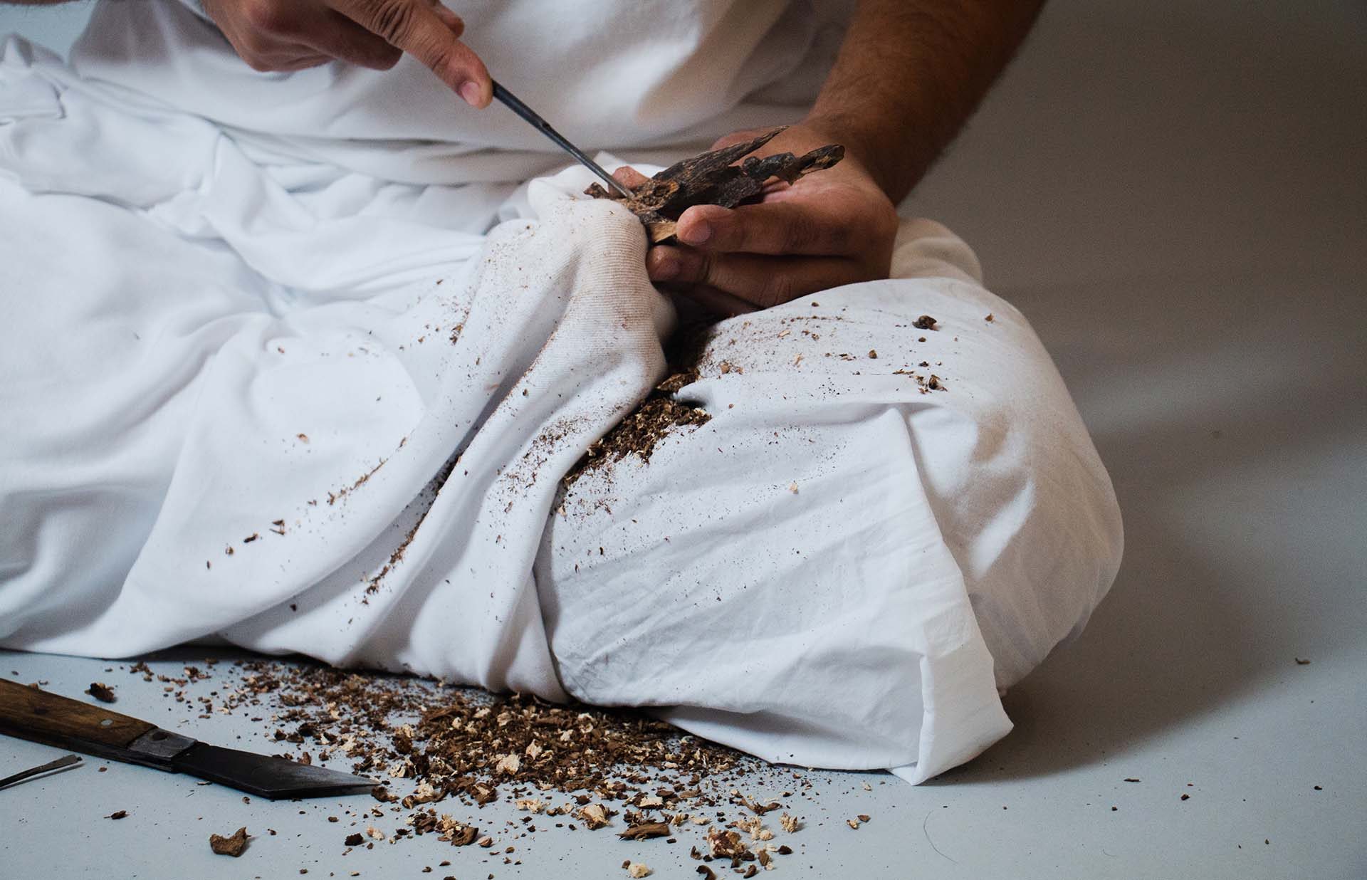 Hands carving agarwood over a white cloth, shavings on the floor
