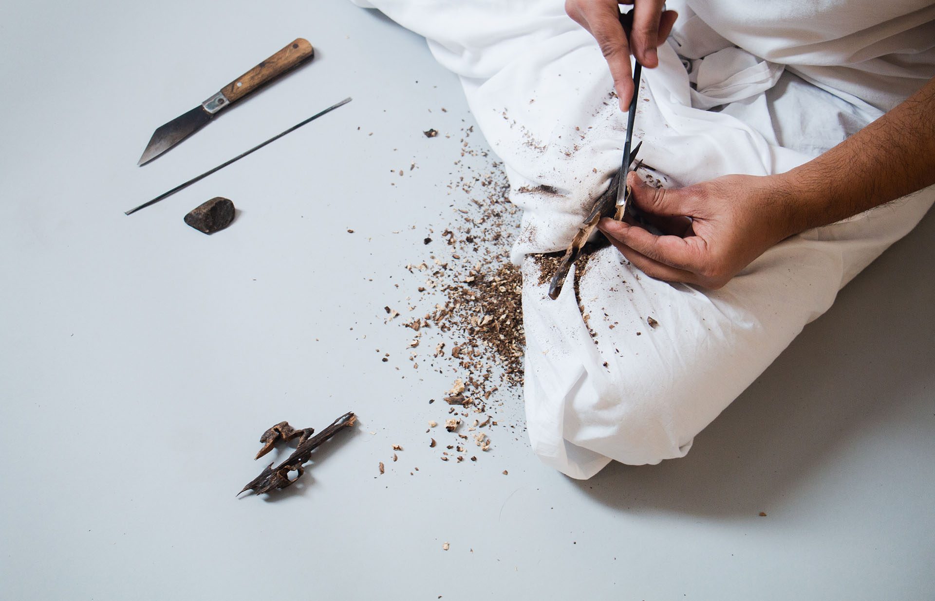 Hands working agarwood on white cloth, a knife and a pick beside them
