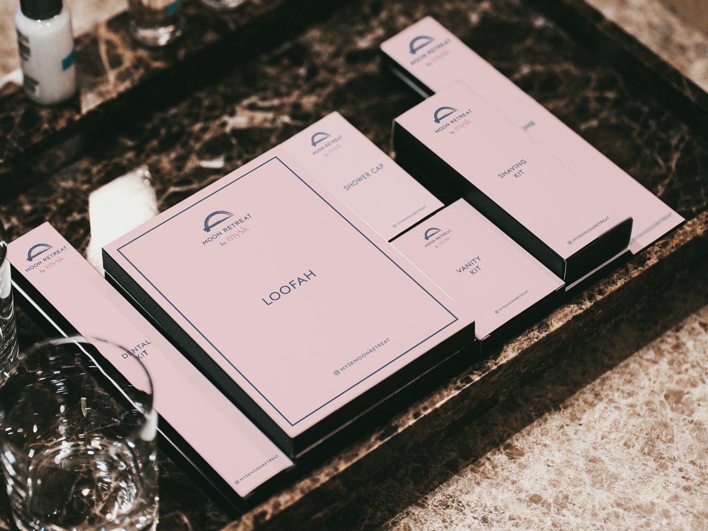 Pink amenity boxes on a dark marble tray: loofah, shower cap, vanity kit
