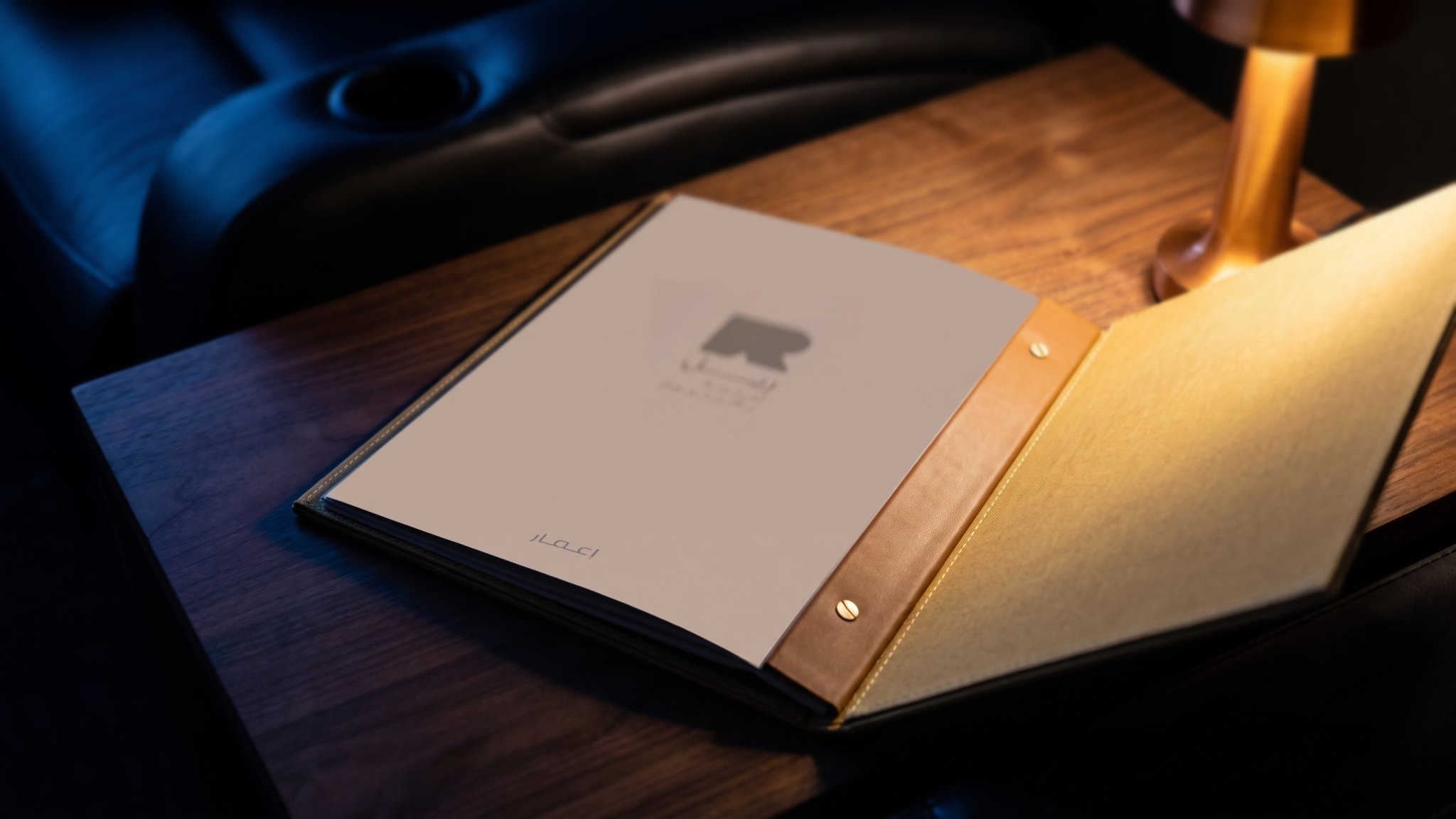 A render: the menu open on a walnut table, its taupe inner cover printed in tone, the leather strap and the gold linen cover beside it