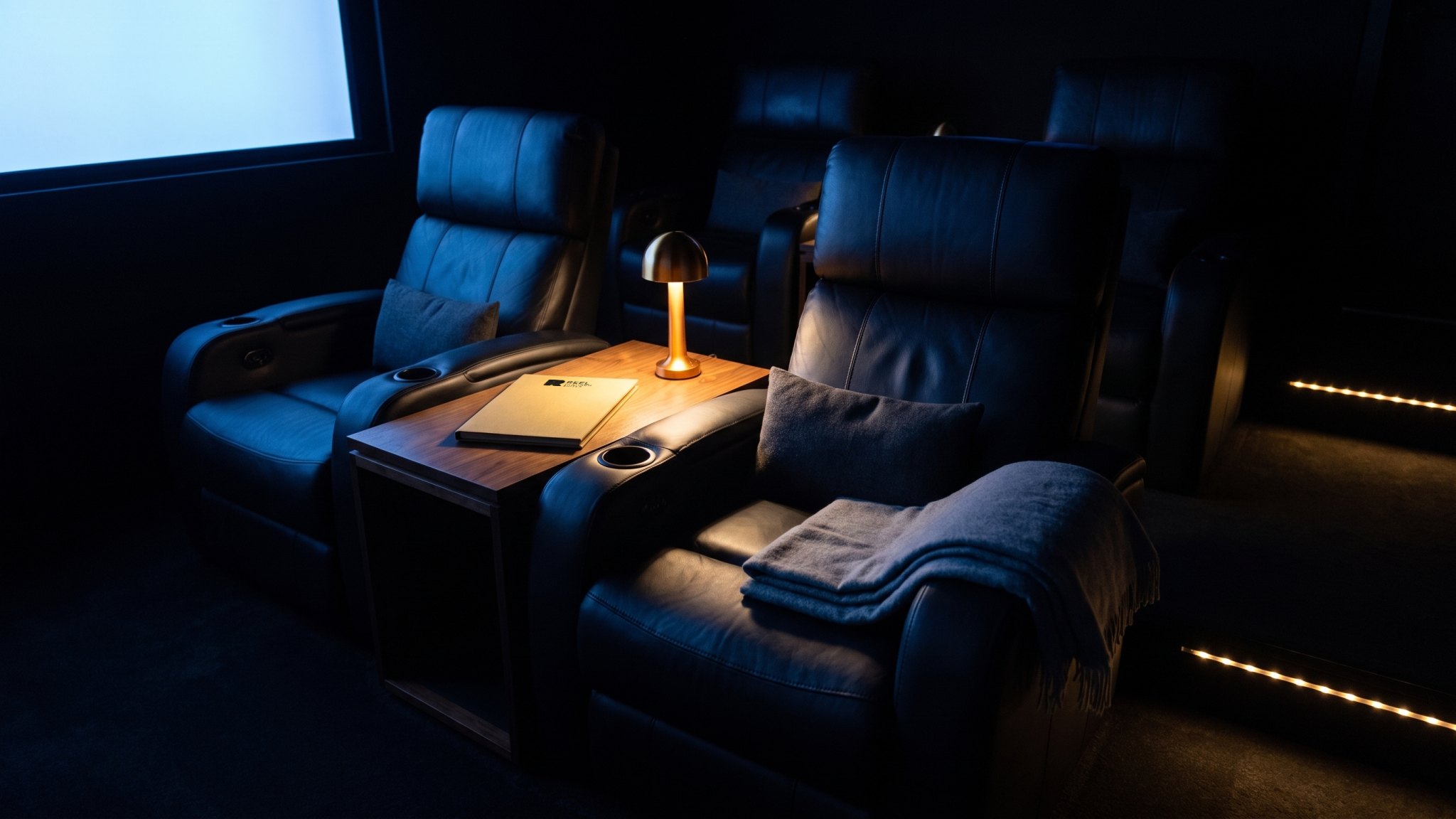 A render: two leather recliners in a dark cinema suite, a lamp and the gold menu on the side table between them