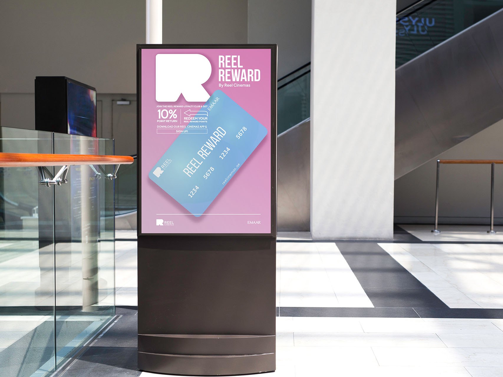A freestanding digital screen in a mall showing the pink Reel Reward poster with a cashback card