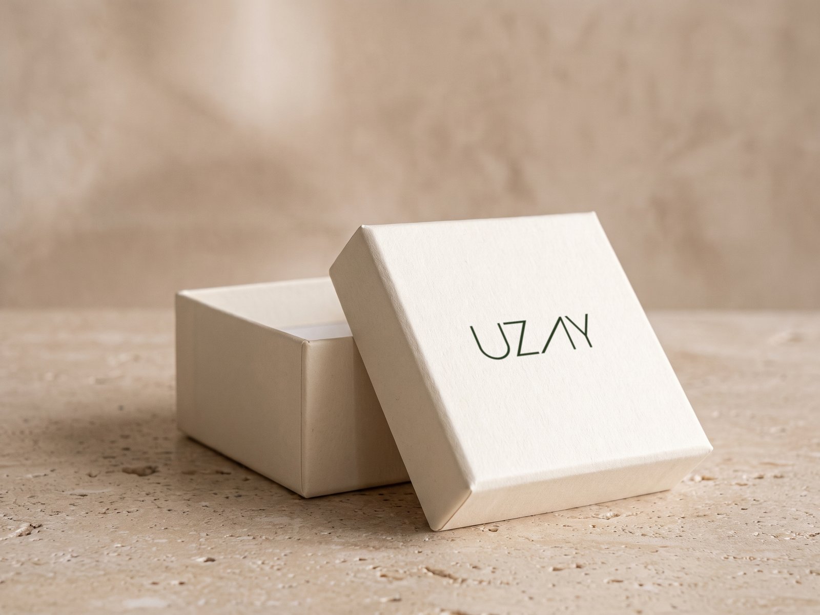 A render: an ivory jewellery box with its lid lifted, the Uzay wordmark on the lid, on travertine