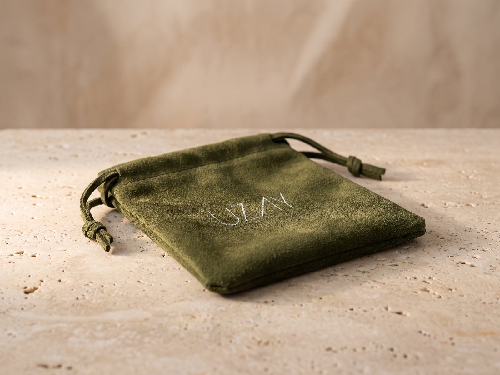 A render: an olive suede jewellery pouch with a drawstring and the Uzay wordmark in white